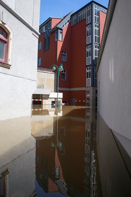 The image depicts a flooded urban alleyway situated between two contrasting buildings, with the floodwater covering the entire ground level and reflecting the surroundings, including a multi-storey mo