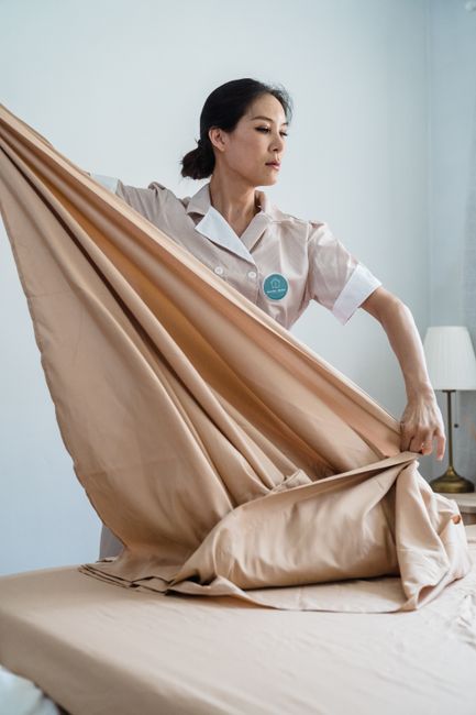 A woman wearing a white uniform with a blue circular badge is in a room, carefully folding or arranging a large beige bedspread or curtain fabric. The fabric appears smooth, with a slight sheen, and i