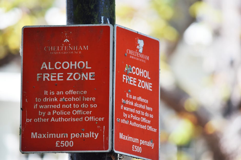 A close-up view of two red metal signboards affixed to a dark metal pole in an outdoor setting, with a blurred background of trees and sunlight. The signs display white text indicating the Cheltenham